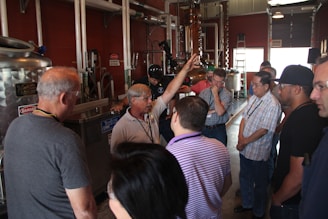 A group of people is gathered around a man gesturing with his hand, likely explaining or demonstrating something in a facility with industrial equipment. The environment seems to be a workshop or a factory setting, with metallic pipes and tanks visible.