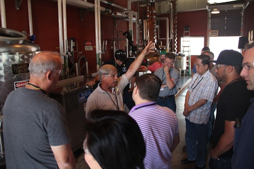 A group of people is gathered around a man gesturing with his hand, likely explaining or demonstrating something in a facility with industrial equipment. The environment seems to be a workshop or a factory setting, with metallic pipes and tanks visible.