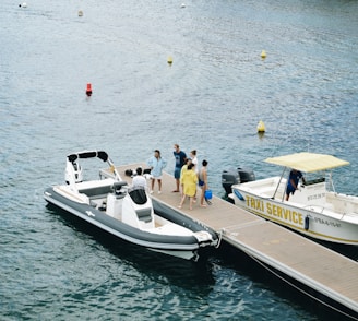 A group of people are gathered on a dock next to two boats in a body of water. One of the boats is a modern white motorboat with black accents, and it is moored to the dock. The other boat, labeled 'Taxi Service,' has a yellow canopy and white hull. The people, dressed in casual summer attire, appear to be preparing to board or disembark. The background features calm waters with some floating buoys.