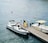 A group of people are gathered on a dock next to two boats in a body of water. One of the boats is a modern white motorboat with black accents, and it is moored to the dock. The other boat, labeled 'Taxi Service,' has a yellow canopy and white hull. The people, dressed in casual summer attire, appear to be preparing to board or disembark. The background features calm waters with some floating buoys.