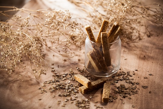 A glass jar filled with rectangular, brown sticks, possibly sesame bars or a similar snack, placed on a wooden surface. Surrounding the jar are scattered seeds and delicate, dried botanical elements, adding a rustic aesthetic.
