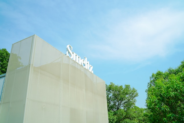 A modern building with a large, translucent facade is depicted. It has a sign on top with white lettering. The surroundings include green trees and a bright blue sky with wispy clouds.