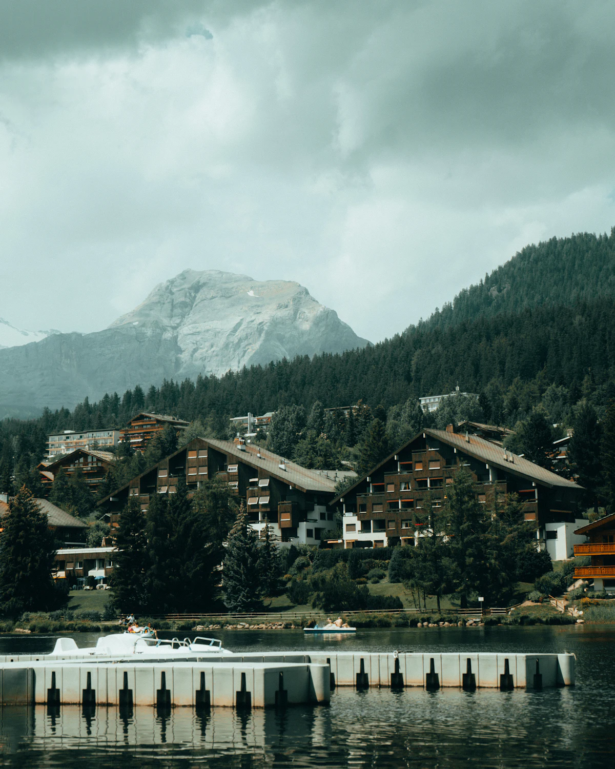 Chalets and dark pines along a lake in Crans-Montana, with a pale rock peak behind under cloud