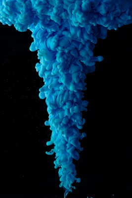 Vibrant blue ink creates swirling, cloud-like formations in water against a stark black background. The intricate patterns resemble a descending plume.