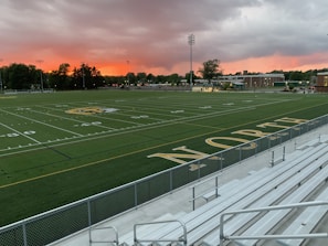 An empty sports field with the word 'NORTH' displayed near the bleachers. The field is well-marked with yard lines and a logo at the center. The sky is covered with dramatic clouds, displaying a vibrant orange and pink hue near the horizon. Trees and buildings surround the field in the distance.