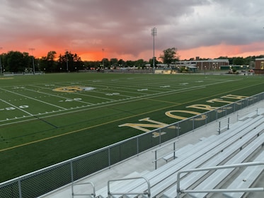 An empty sports field with the word 'NORTH' displayed near the bleachers. The field is well-marked with yard lines and a logo at the center. The sky is covered with dramatic clouds, displaying a vibrant orange and pink hue near the horizon. Trees and buildings surround the field in the distance.