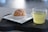 A cream puff sits on a small white square plate, placed on a dark wooden board. Next to it is a transparent glass cup filled with a light greenish-yellow liquid, possibly tea.