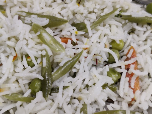 A close-up view of a dish consisting of basmati rice mixed with vegetables. The white rice grains are interspersed with vibrant green beans, peas, and orange carrot slices, suggesting a vegetable pulao or biryani. The texture of the rice appears fluffy and slightly glistening.
