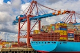 A large cargo ship is docked at a port, stacked high with multicolored shipping containers. An industrial crane towers above the ship, indicating active loading or unloading. The sky is partly cloudy, and there are wind turbines visible in the background, suggesting a modern port environment.