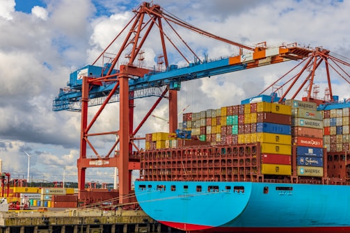 A large cargo ship is docked at a port, stacked high with multicolored shipping containers. An industrial crane towers above the ship, indicating active loading or unloading. The sky is partly cloudy, and there are wind turbines visible in the background, suggesting a modern port environment.