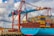 A large cargo ship is docked at a port, stacked high with multicolored shipping containers. An industrial crane towers above the ship, indicating active loading or unloading. The sky is partly cloudy, and there are wind turbines visible in the background, suggesting a modern port environment.