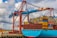 A large cargo ship is docked at a port, stacked high with multicolored shipping containers. An industrial crane towers above the ship, indicating active loading or unloading. The sky is partly cloudy, and there are wind turbines visible in the background, suggesting a modern port environment.