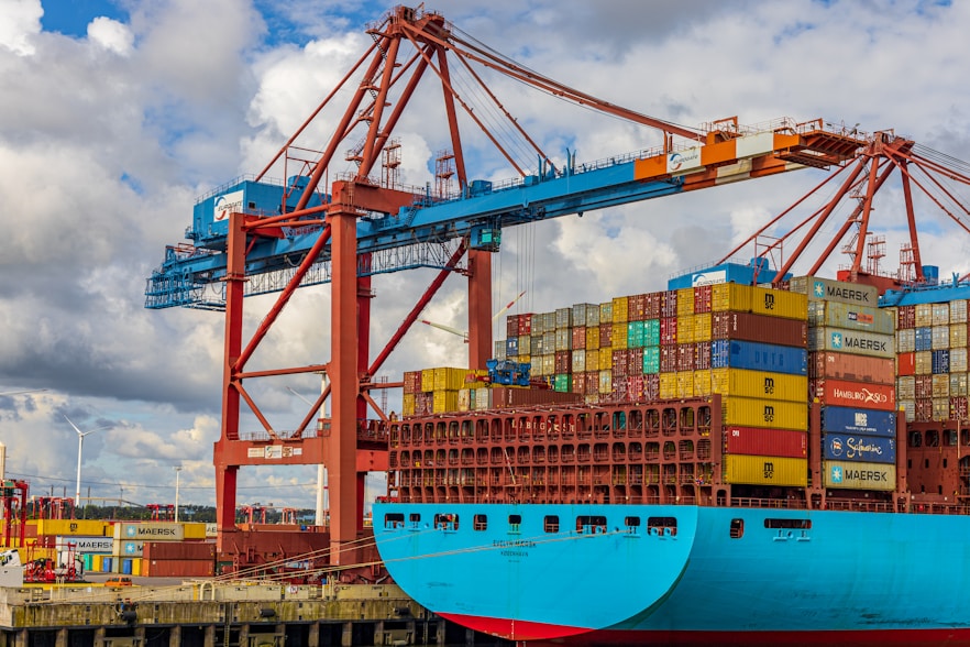 A large cargo ship is docked at a port, stacked high with multicolored shipping containers. An industrial crane towers above the ship, indicating active loading or unloading. The sky is partly cloudy, and there are wind turbines visible in the background, suggesting a modern port environment.