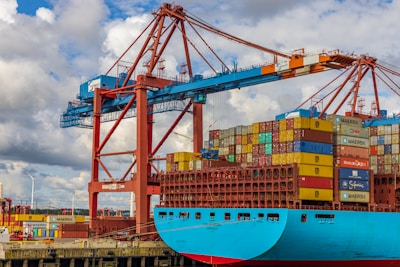 A large cargo ship is docked at a port, stacked high with multicolored shipping containers. An industrial crane towers above the ship, indicating active loading or unloading. The sky is partly cloudy, and there are wind turbines visible in the background, suggesting a modern port environment.