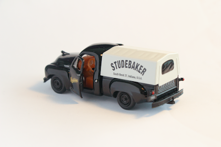 A miniature model of a vintage Studebaker truck with an open driver's side door. The truck has a black cab and a white canopy cover with 'STUDEBAKER' printed on the side.