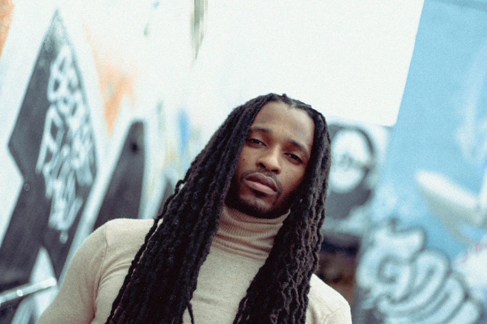 Man with dreadlocks stands against graffiti-covered backdrop with direct gaze towards the viewer