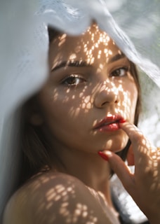 A woman is partially obscured by a lace curtain that casts intricate shadows across her face. Her eyes gaze directly at the viewer, and her fingers with red nail polish lightly touch her lips. The lighting creates a soft, intimate atmosphere.