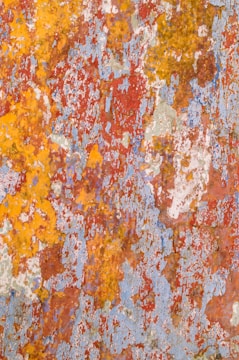 The image displays a surface with peeling and flaking paint, revealing various layers of color. The colors include shades of red, yellow, blue, white, and some rust. The texture is rough, and the paint appears aged and weathered.