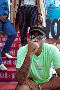 A person wearing a bright green T-shirt with 'Positive Vibes' written on it, adorned with tattoos on their arm and a chain necklace with a pendant. They are seated on red stairs that feature white graffiti text. Two other individuals stand behind, one dressed in a blue suit with sparkling shoes and the other in striped trousers and a beige coat. The background consists of colorful graffiti.
