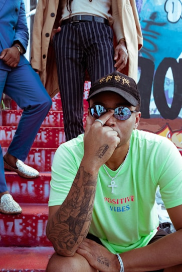 A person wearing a bright green T-shirt with 'Positive Vibes' written on it, adorned with tattoos on their arm and a chain necklace with a pendant. They are seated on red stairs that feature white graffiti text. Two other individuals stand behind, one dressed in a blue suit with sparkling shoes and the other in striped trousers and a beige coat. The background consists of colorful graffiti.