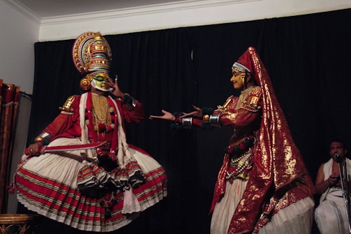 Two performers in traditional Indian dance costumes are engaged in a dramatic pose. The left performer wears an elaborate headpiece, colorful face paint, and a voluminous skirt, while striking a theatrical gesture. The right performer, adorned with a sequined head covering and a richly decorated outfit, extends their hands towards the other performer. A musician in the background adds to the ambiance.