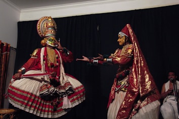 Two performers in traditional Indian dance costumes are engaged in a dramatic pose. The left performer wears an elaborate headpiece, colorful face paint, and a voluminous skirt, while striking a theatrical gesture. The right performer, adorned with a sequined head covering and a richly decorated outfit, extends their hands towards the other performer. A musician in the background adds to the ambiance.