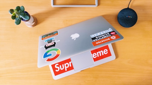 A wooden table holds a closed laptop adorned with various colorful stickers, including brand names and logos. A small potted plant with green leaves is situated to the left, while a round, black smart speaker with colored lights sits on the right.