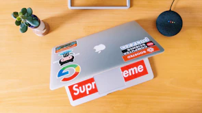 An open laptop on a wooden desk with colorful stickers, surrounded by coffee and tech magazines.