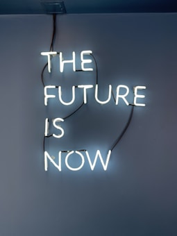 A neon sign with the phrase 'THE FUTURE IS NOW' mounted on a dark blue wall. The letters are glowing in a bright white light, with visible wires connecting them.