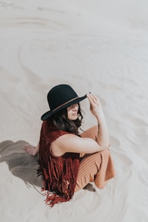 A person sitting on sand, wearing a black wide-brimmed hat and a sleeveless, fringed vest. The sand is light in color and the sky is clear.