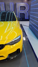 A vibrant yellow car is parked indoors, partially seen from the front side. The design is sleek with a focus on aerodynamic features and modern headlight styling. In the background, large letters form a decorative element on the wall, highlighted by blue neon lighting. The setting includes a futuristic ambiance with reflective surfaces and blue accent lighting.