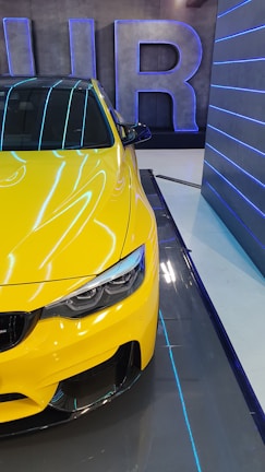 A vibrant yellow car is parked indoors, partially seen from the front side. The design is sleek with a focus on aerodynamic features and modern headlight styling. In the background, large letters form a decorative element on the wall, highlighted by blue neon lighting. The setting includes a futuristic ambiance with reflective surfaces and blue accent lighting.