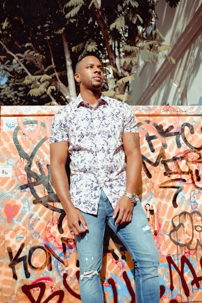 A man stands confidently against a colorful graffiti-covered wall. He wears a floral-patterned short-sleeve shirt and ripped jeans. Sunlight filters through the trees above, creating shadows on the wall.