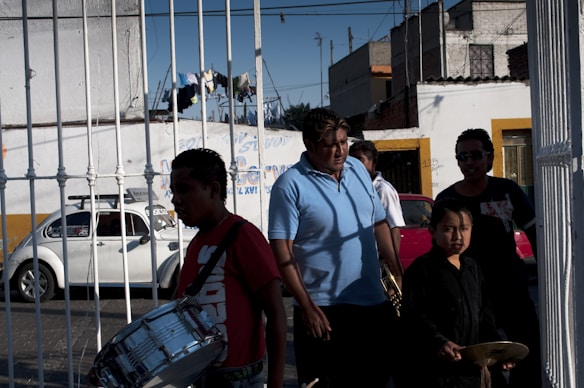 A group of people, including a child, appear to be walking along a street. Some of them are holding musical instruments, such as a drum and cymbals. There is a white car parked near a building in the background, and laundry is seen hanging above the scene. The walls of the buildings are painted in neutral colors with some yellow and white details.