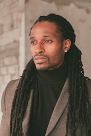 A person with long dreadlocks wears a dark turtleneck and a gray blazer, gazing thoughtfully to the side. The backdrop appears to be a textured indoor setting with neutral tones.