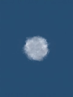 A single fluffy cloud is set against a deep blue sky, centered in the image. The cloud appears soft and slightly translucent with subtle shading indicating light and shadow.