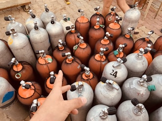 Several scuba tanks, some with orange netting and other metallic grey ones, are clustered together on a tiled floor. A person's hand reaches out towards one of the tanks, and another person's legs are partially visible in the background.