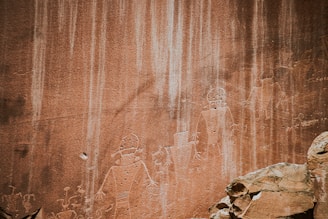 Ancient petroglyphs etched into a sandstone rock face, depicting humanoid figures with distinct outlines and some intricate patterns. The rock has natural striations and varying shades of earthy tones, with boulders or stones visible in the foreground.