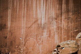 Ancient petroglyphs etched into a sandstone rock face, depicting humanoid figures with distinct outlines and some intricate patterns. The rock has natural striations and varying shades of earthy tones, with boulders or stones visible in the foreground.