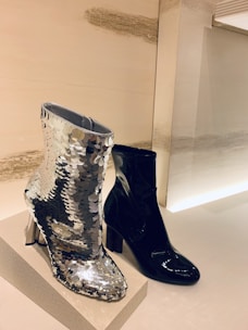Two ankle boots are on display, one covered in silver sequins that reflect the light, and the other in glossy black material. They are placed on a textured platform against a minimalist background.