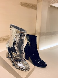 Two ankle boots are on display, one covered in silver sequins that reflect the light, and the other in glossy black material. They are placed on a textured platform against a minimalist background.