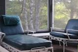 A cozy room featuring a blue cushioned reclining chair and armchair with white metal frames. The chairs are positioned alongside a small white table. Large windows provide a view of lush green trees outside, creating a serene atmosphere.