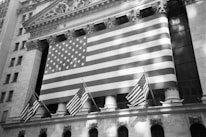 A large American flag hangs prominently on the exterior of a classical-style building, which has ornate architectural details and pillars. Smaller American flags are also displayed, adding to the patriotic theme.