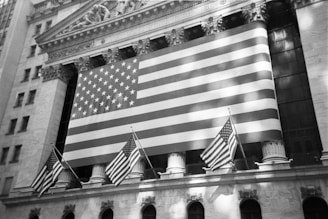 A large American flag hangs prominently on the exterior of a classical-style building, which has ornate architectural details and pillars. Smaller American flags are also displayed, adding to the patriotic theme.