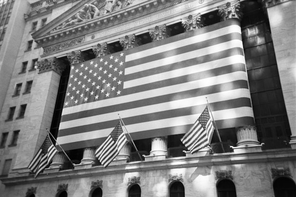 A large American flag hangs prominently on the exterior of a classical-style building, which has ornate architectural details and pillars. Smaller American flags are also displayed, adding to the patriotic theme.