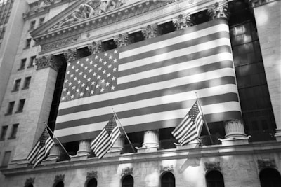 A large American flag hangs prominently on the exterior of a classical-style building, which has ornate architectural details and pillars. Smaller American flags are also displayed, adding to the patriotic theme.