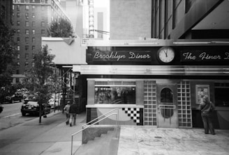 A street scene with a classic diner featuring a sign that reads 'Brooklyn Diner' and an analog clock showing 2:00. The diner has a retro design with checkered patterns. To the right, a person stands near the entrance while two people walk on the sidewalk nearby. Tall buildings and a parked car are visible in the background.