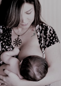 A woman is breastfeeding a baby, holding the child gently in her arms. Her gaze is directed downwards, and she wears a patterned outfit with a large pendant necklace. The scene appears intimate and nurturing.