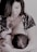 A woman is breastfeeding a baby, holding the child gently in her arms. Her gaze is directed downwards, and she wears a patterned outfit with a large pendant necklace. The scene appears intimate and nurturing.