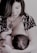 A woman is breastfeeding a baby, holding the child gently in her arms. Her gaze is directed downwards, and she wears a patterned outfit with a large pendant necklace. The scene appears intimate and nurturing.